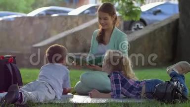 课外活动，可爱的孩子放学后坐在大自然的绿草上听女人读书